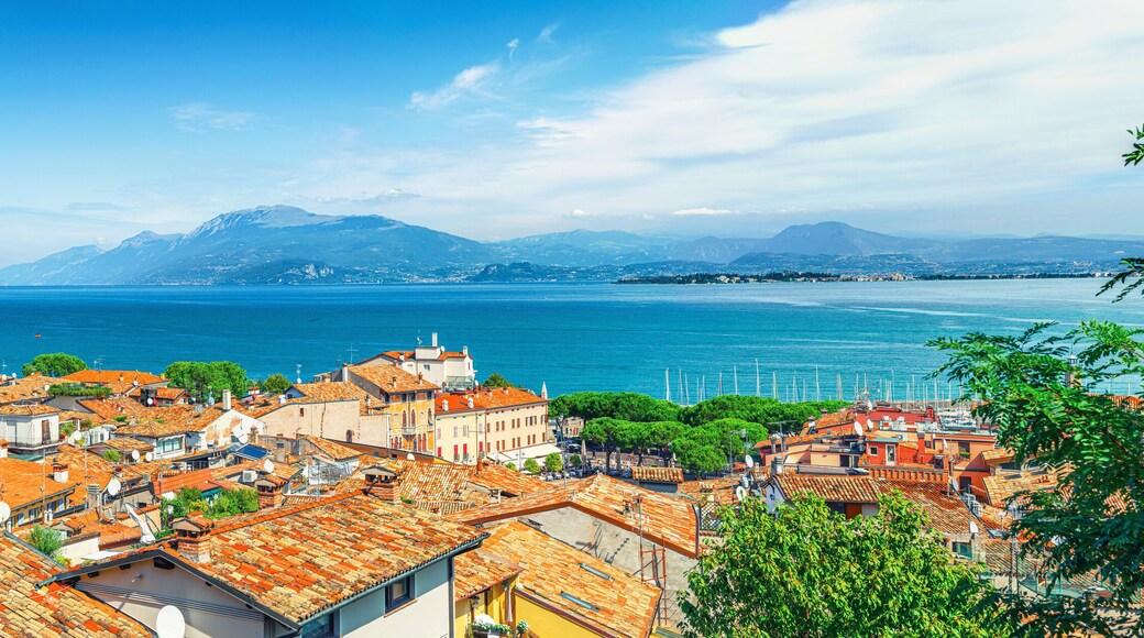 Panorama of Desenzano del Garda town with red tiled roof buildings, Garda Lake water, Monte Baldo mountain range, Sirmione peninsula, Lombardy, Northern Italy. Aerial panoramic view of Desenzano
