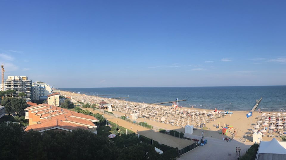 A beach with umbrellas and sun loungers, a pier extending into the water, and buildings in the background.
