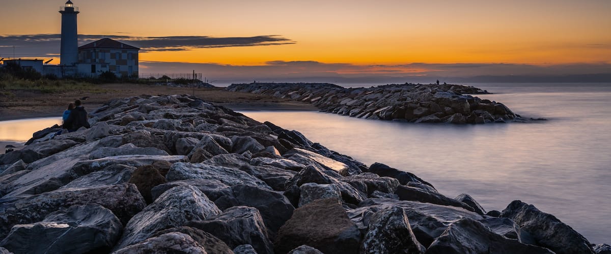 Eine felsige Küstenlinie mit einem Leuchtturm und einem Gebäude, ein ruhiges Meer und ein Sonnenuntergangshimmel.