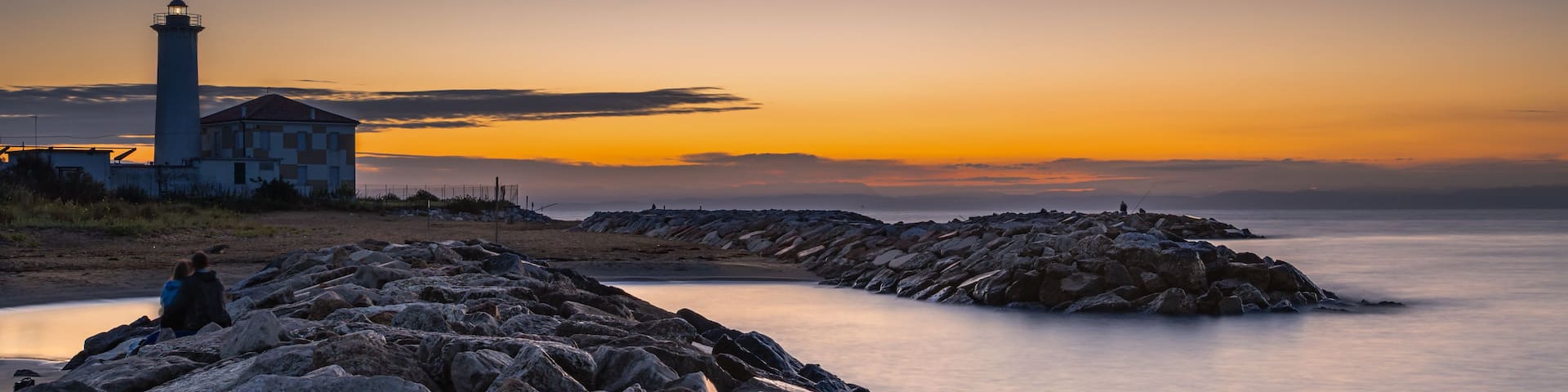 A rocky shoreline with a lighthouse and a building, a calm sea, and a sunset sky.