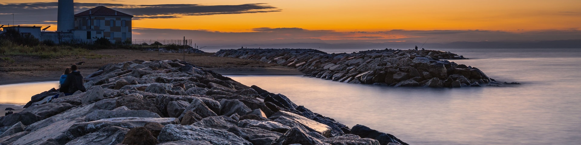 A rocky shoreline with a lighthouse and a building, a calm sea, and a sunset sky.
