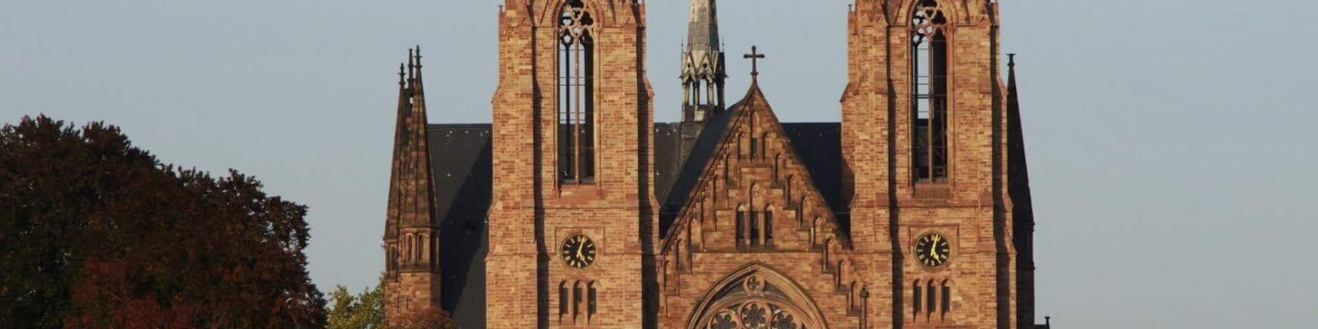 A historic church with tall spires and a large rose window, located beside a river with a boat passing by.