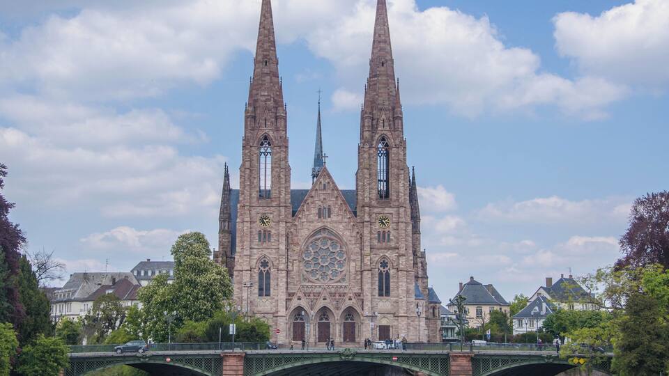 A historic church with twin spires and a large rose window, across a river with a green bridge.