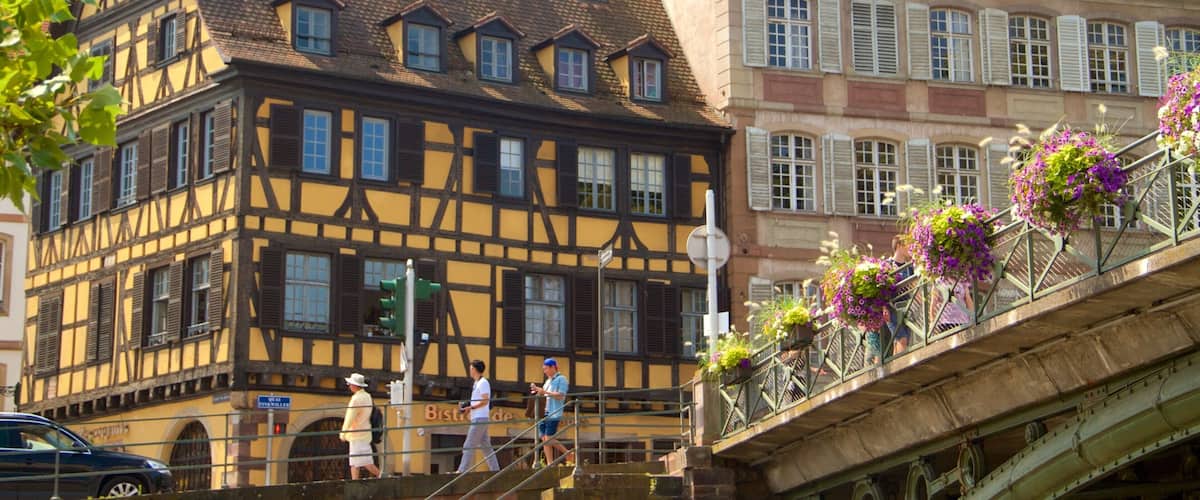 A traditional half-timbered building with a steeply pitched roof and dormer windows, adjacent to a pedestrian bridge with flower baskets.