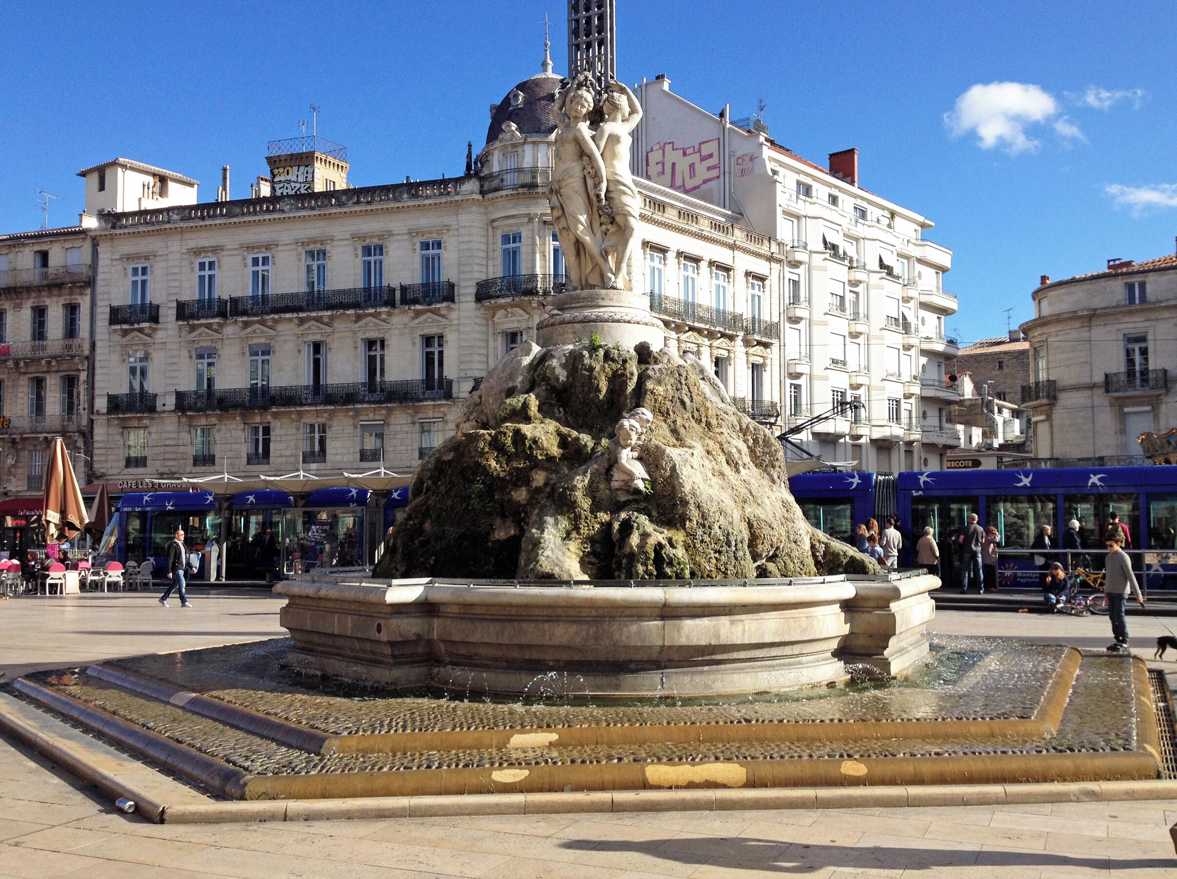 Une fontaine historique ornée d’une statue, entourée d’une place bordée de bâtiments et de cafés.