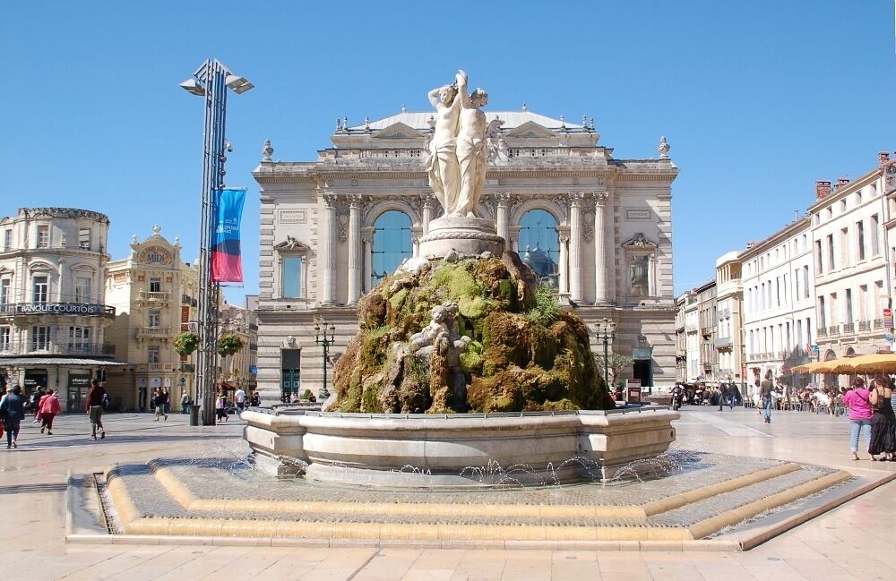 A public square with a central fountain, surrounded by classical architecture and a large building with arched windows.