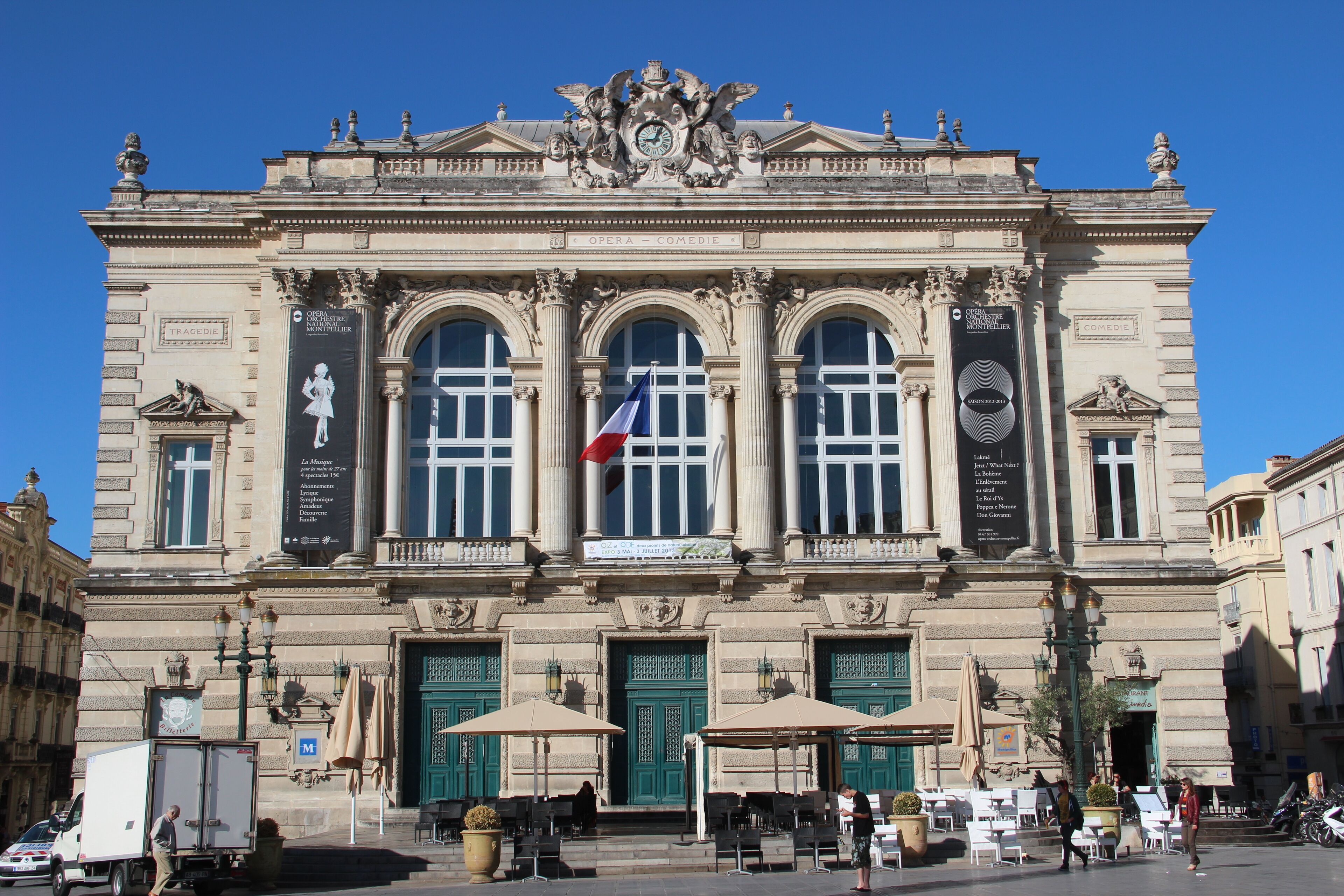 Ein historisches Opernhaus mit aufwendiger Architektur, großen grünen Türen und Außenbereichen mit Sitzgelegenheiten.