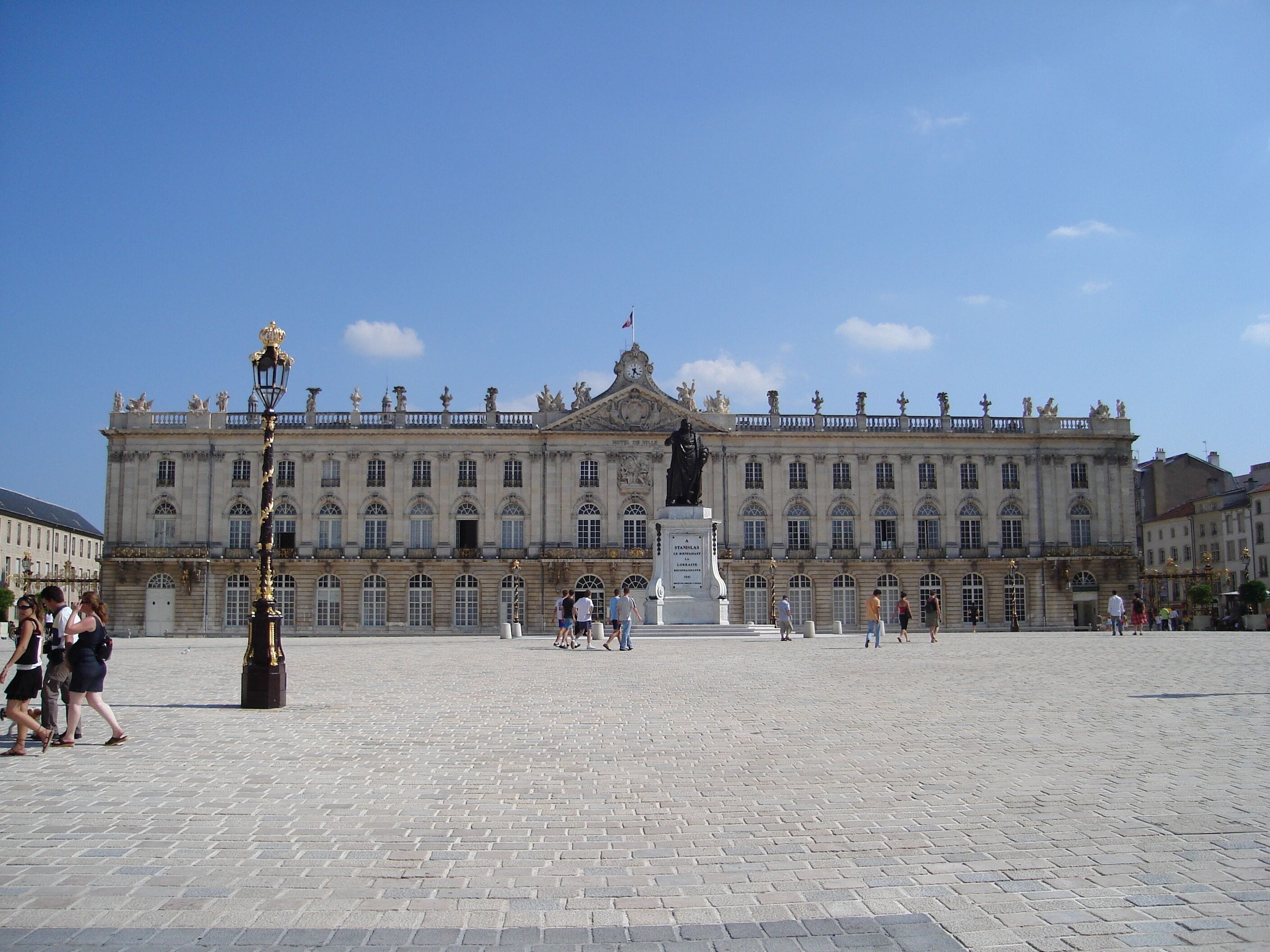 Un grand bâtiment historique, doté d’une statue centrale et bordé d’une place pavée.