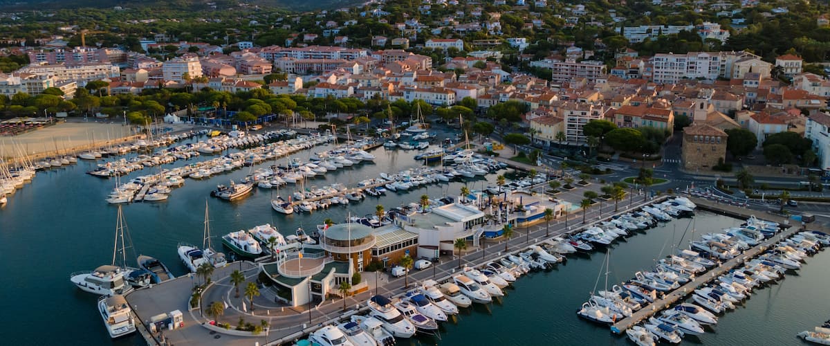 A marina with numerous boats docked, surrounded by a coastal town with buildings and hills in the background.