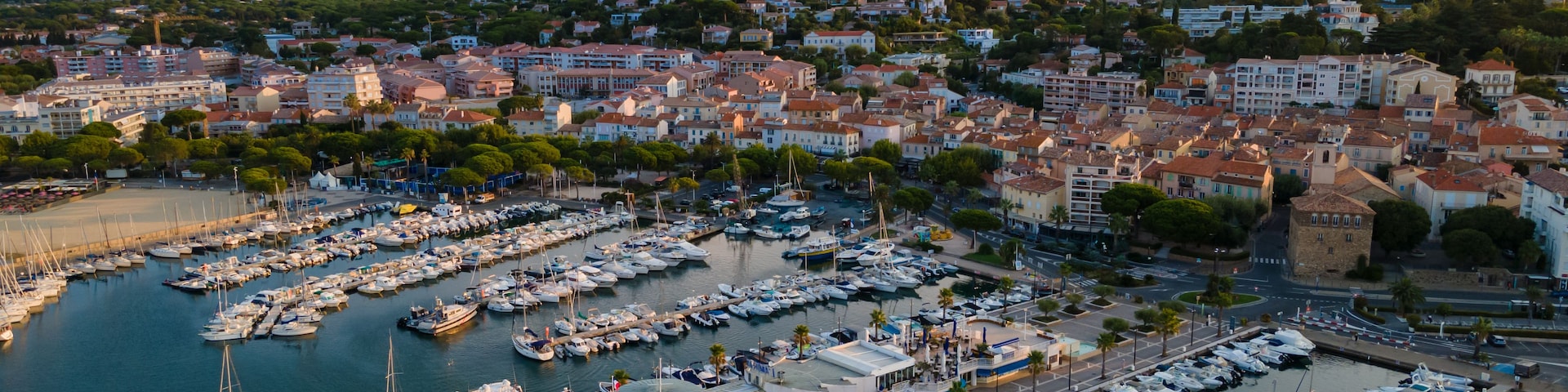 Un puerto deportivo con numerosas embarcaciones amarradas, rodeado por un pueblo costero con edificios y colinas al fondo.