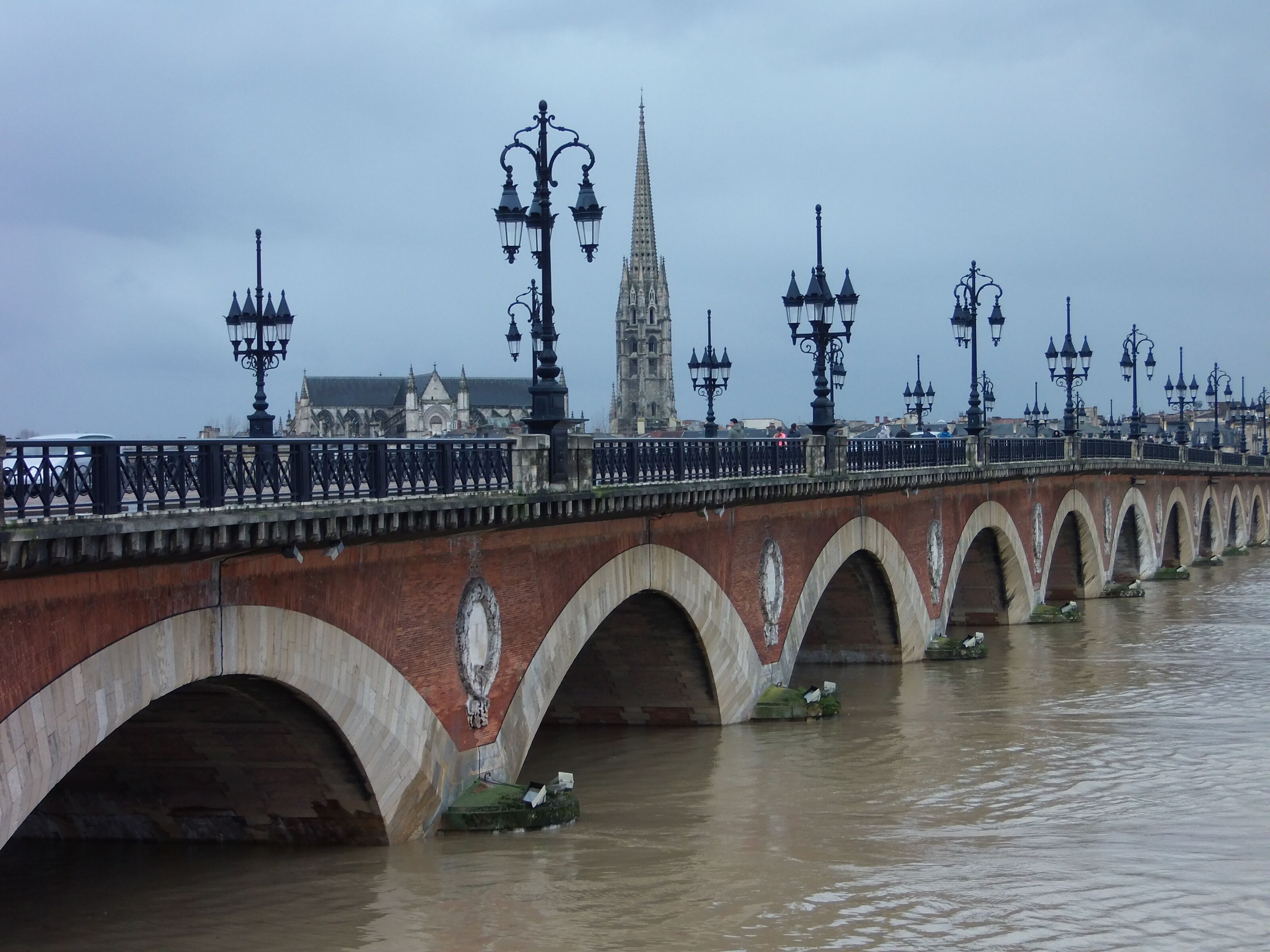 A stone bridge with multiple arches over a river, accompanied by street lamps and a distant church spire.
