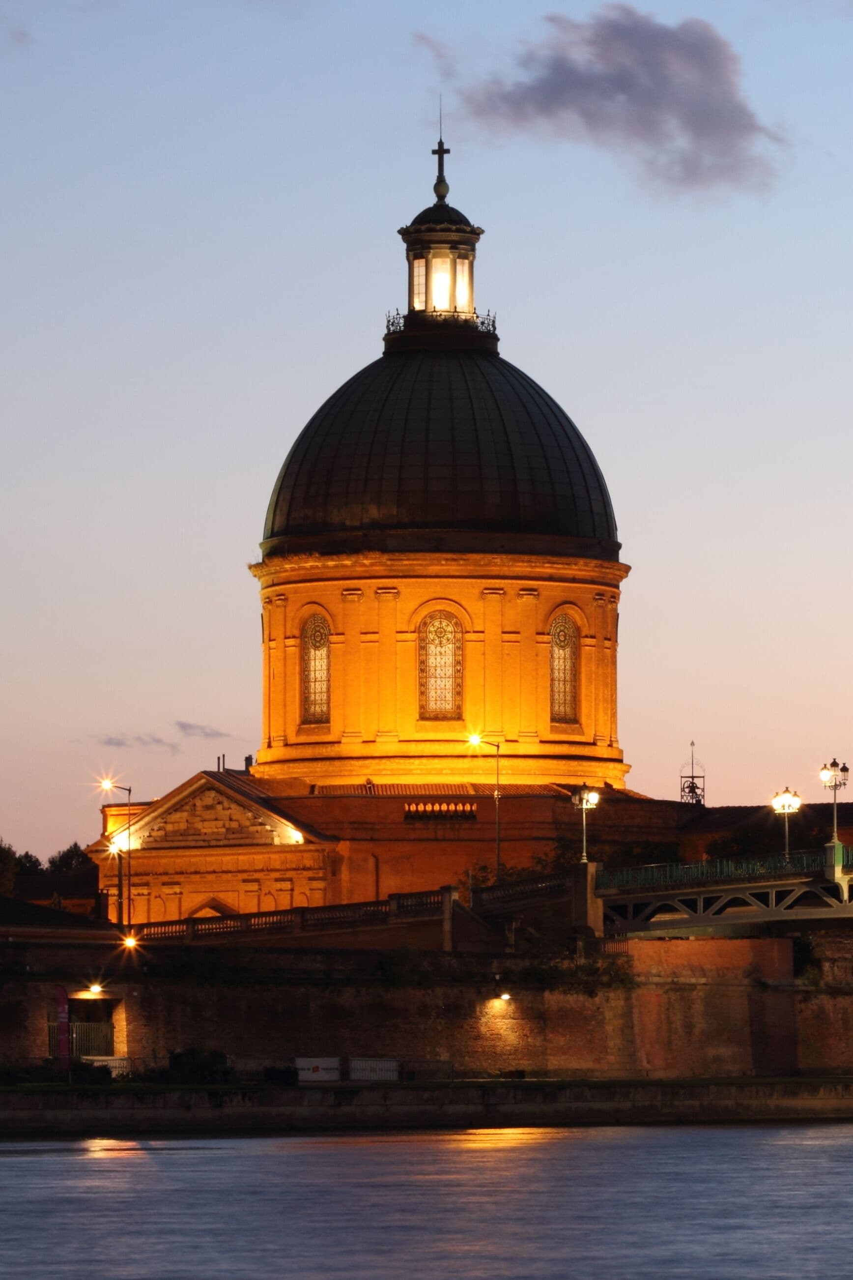 A large domed building with a cross on top, illuminated by warm lights, situated by a body of water.