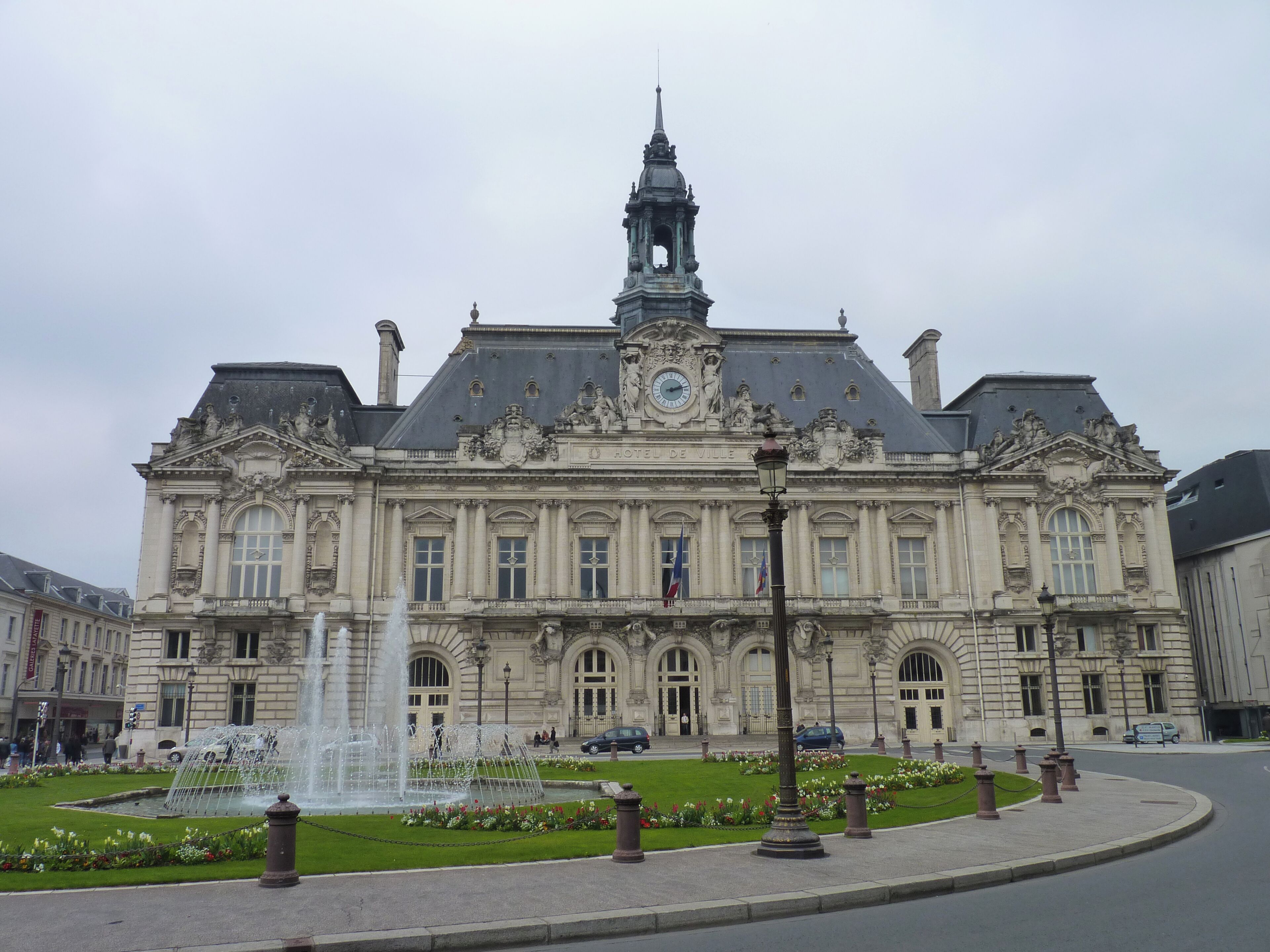 A large historic building with a prominent clock tower and a fountain in front.