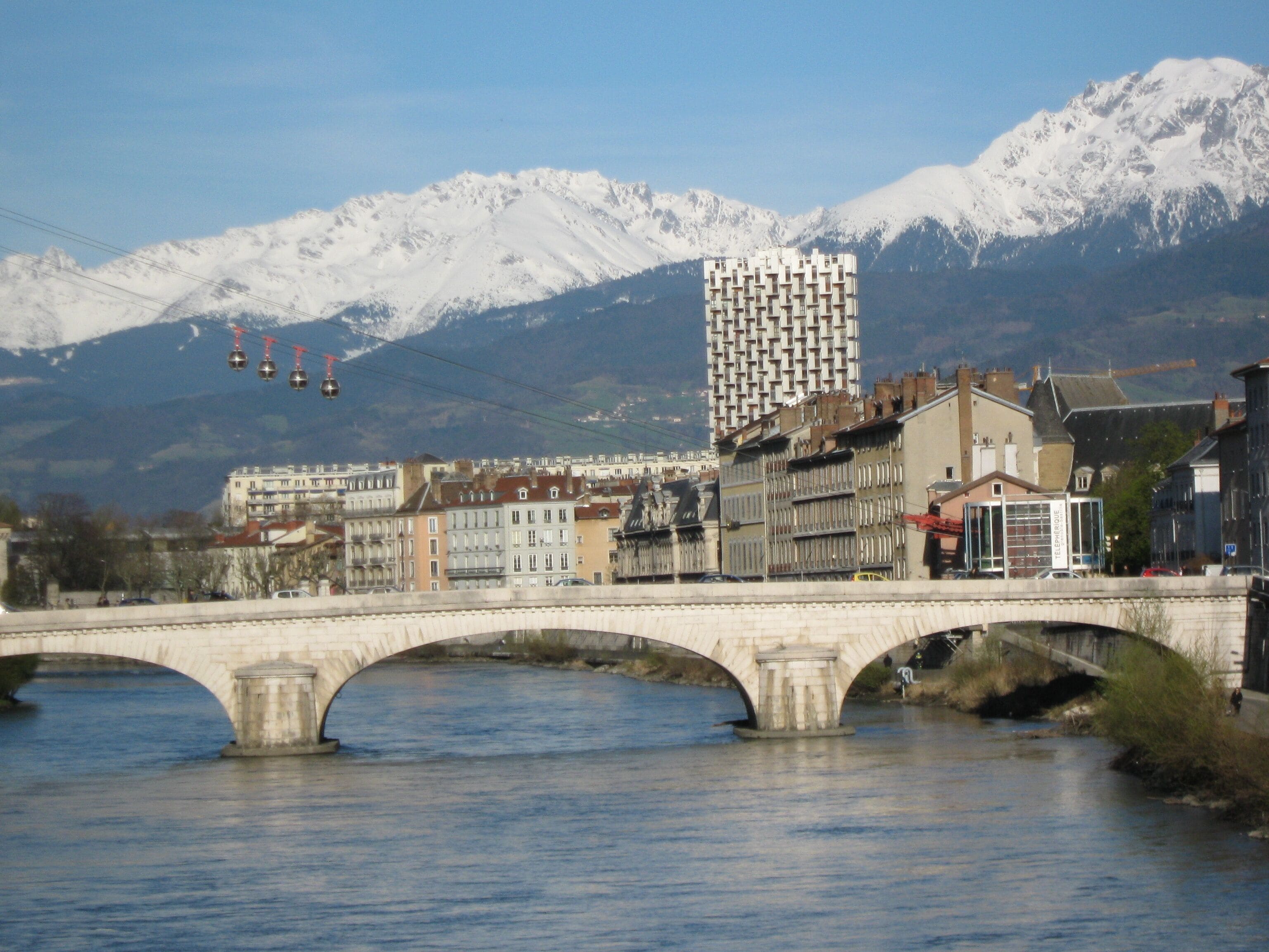 A stone bridge over a river with snow-capped mountains in the background and a modern building with a unique facade.