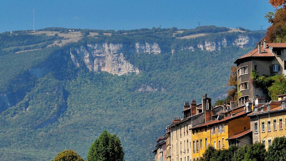 Un téléphérique, un pont de pierre et des bâtiments le long d’une rivière, avec une chaîne de montagnes en arrière-plan.