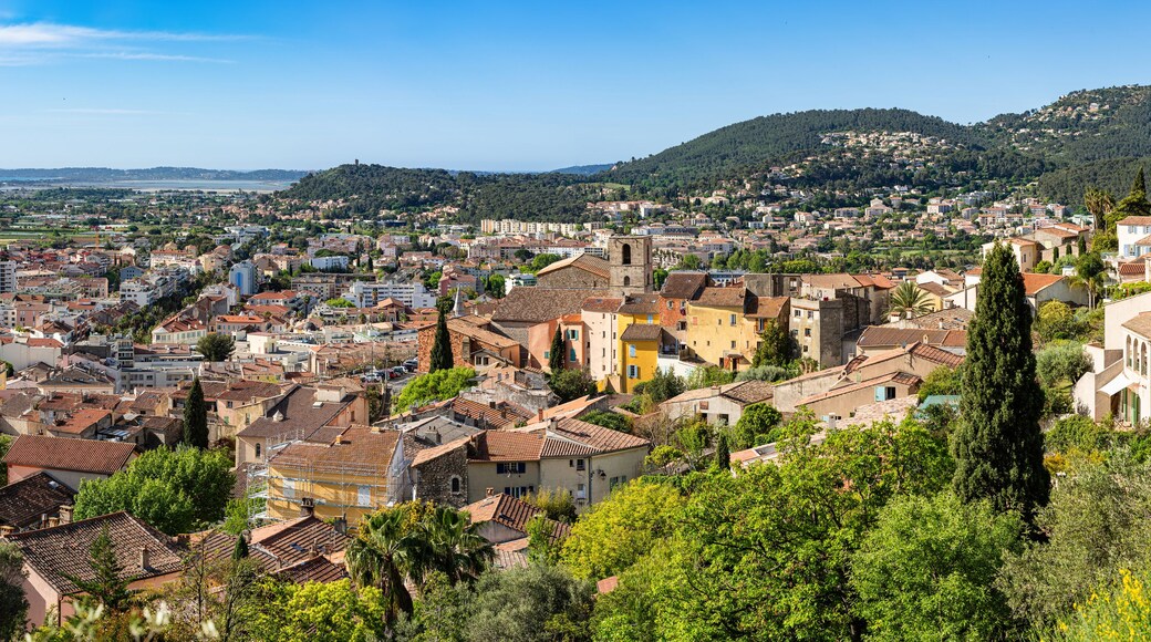 A coastal town with terracotta-roofed buildings, lush greenery, and a mountainous backdrop.