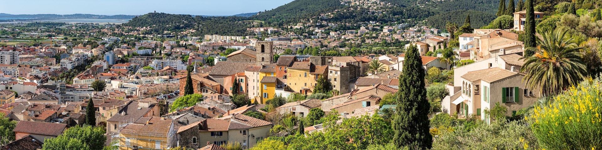 Une ville côtière aux bâtiments aux toits de tuiles rouges, à la végétation luxuriante et dominée par un paysage montagneux.