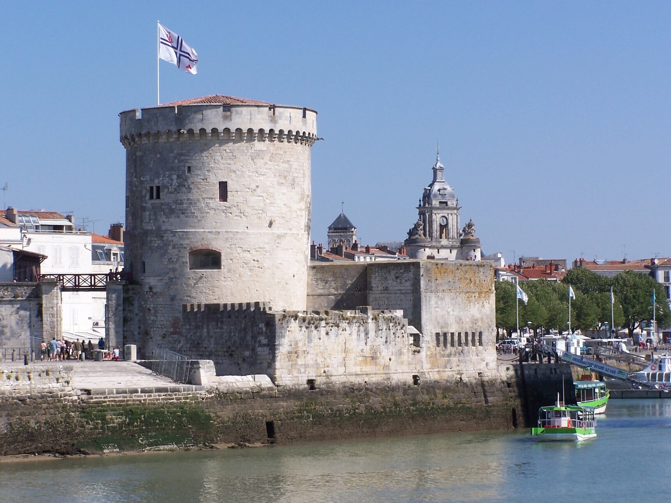 A historic tower with a flag, a church with a dome, and a river with boats.
