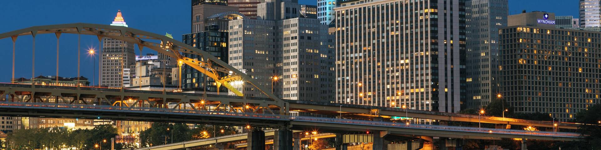 A city skyline at dusk with illuminated buildings, a prominent bridge, and a river.