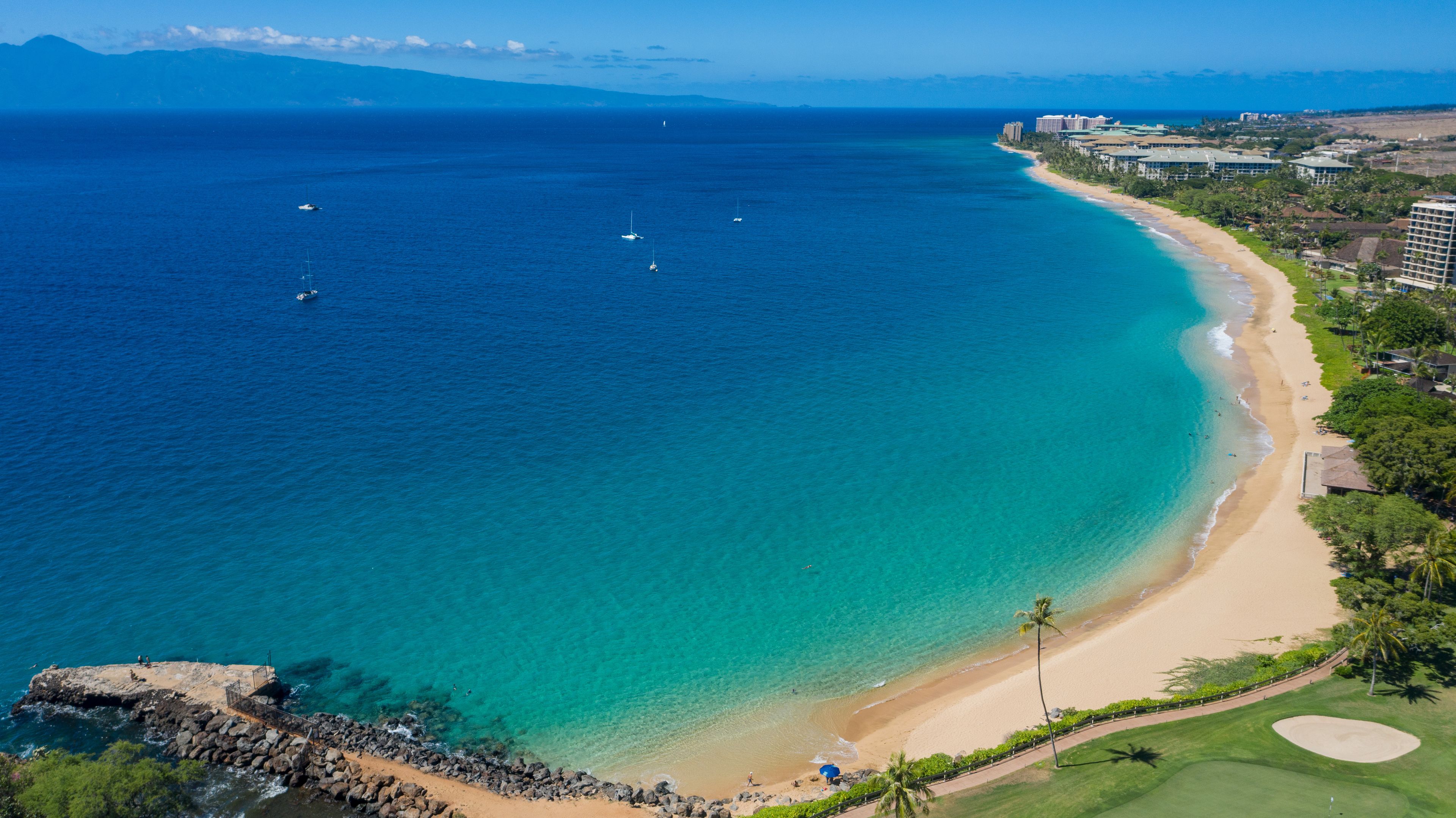 A coastal landscape with a clear blue sea, a sandy beach, and a golf course.