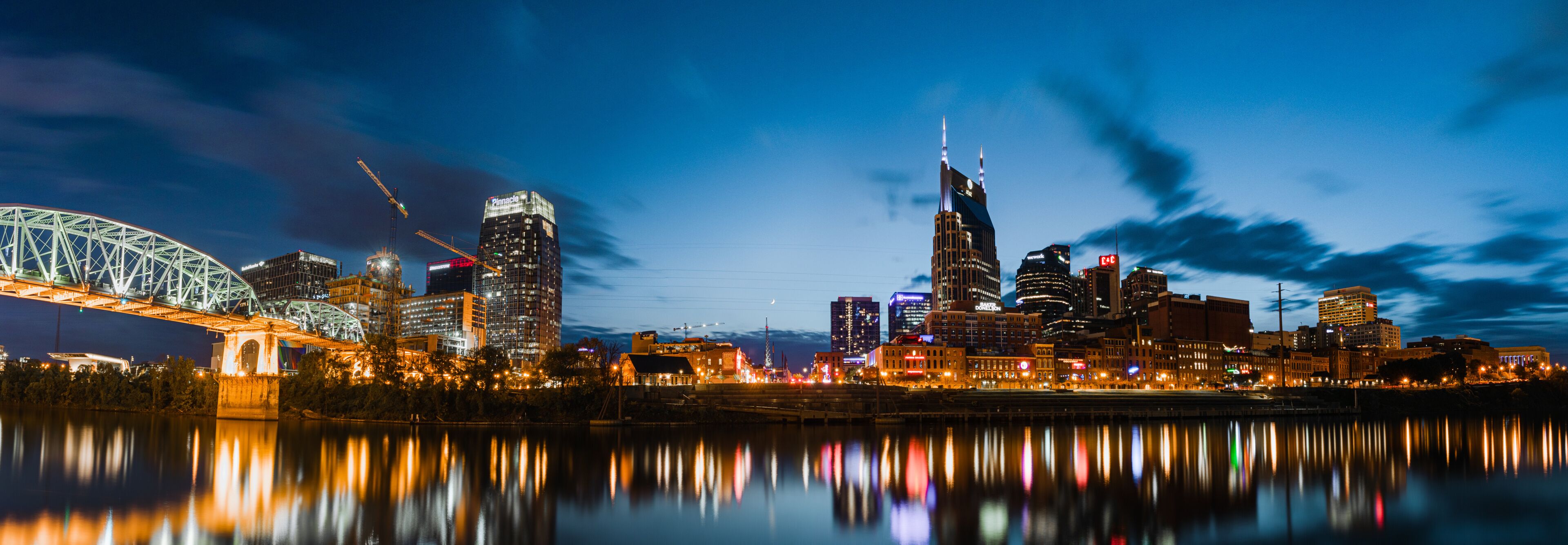 A city skyline at night with a prominent bridge and skyscrapers reflecting on the water.