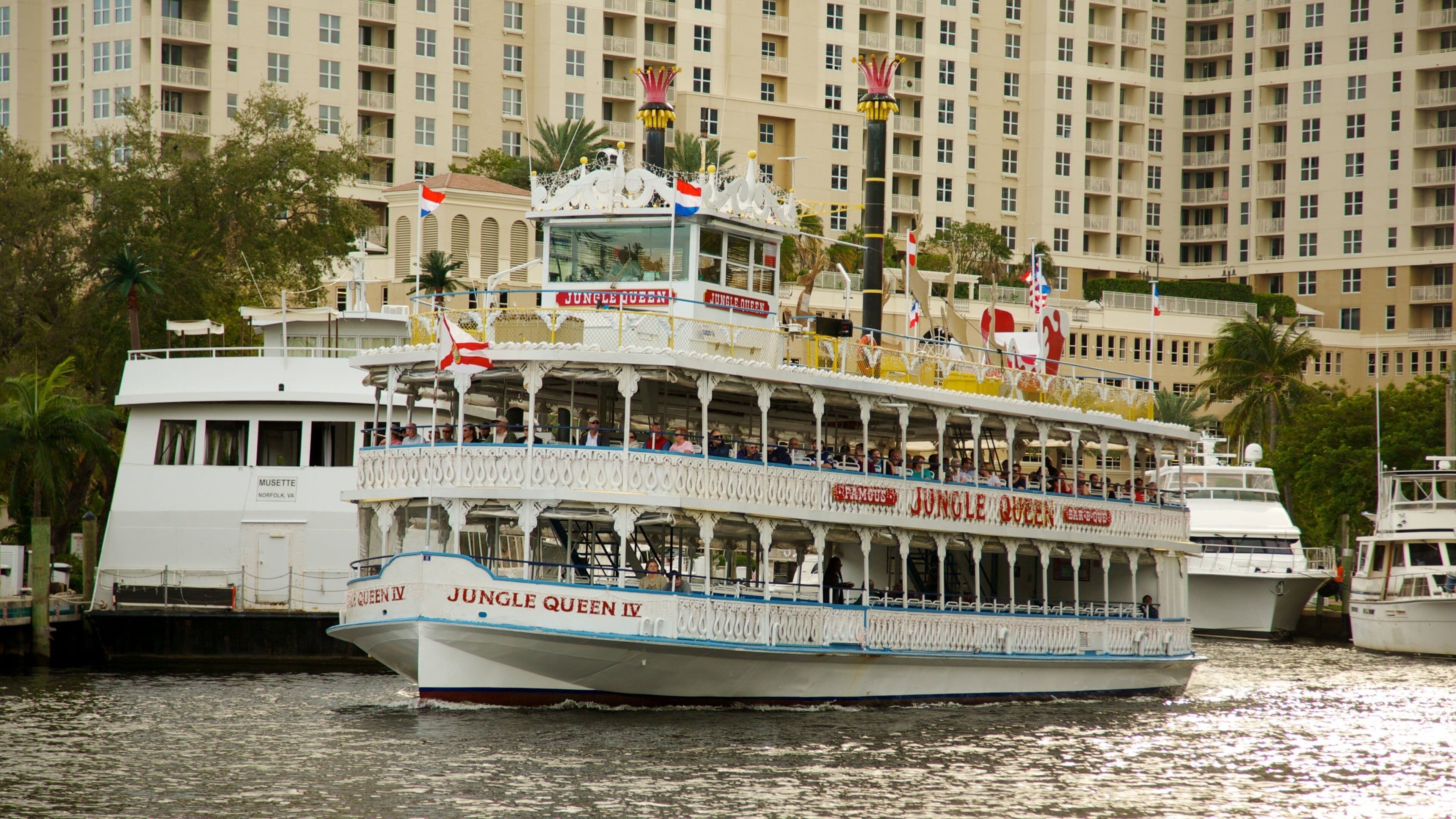A white cruise ship named 'Jungle Queen IV' with multiple decks and flags, docked near a large hotel building.