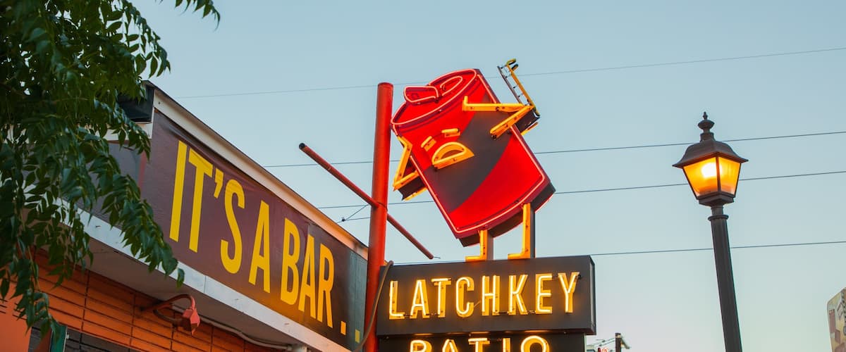 A bar named "It's a Bar" with a neon sign for "Latchkey Patio" and a street lamp.