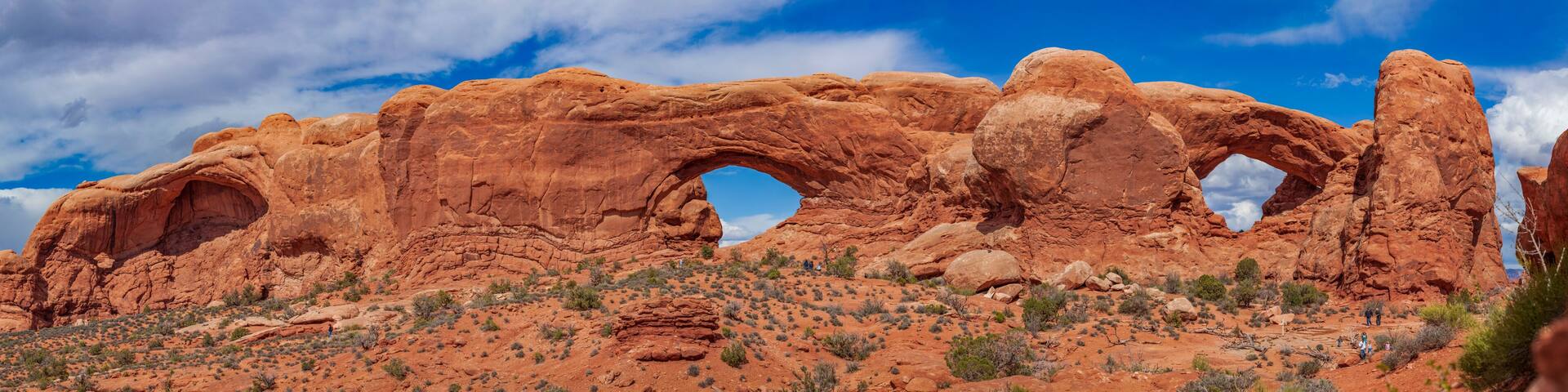 Panoramic view of North Window, South Window, and Turret Arch with beautiful clouds in the sky, small figures of people in the frame emphasize the scale and beauty of this majestic creation, Moab, USA
