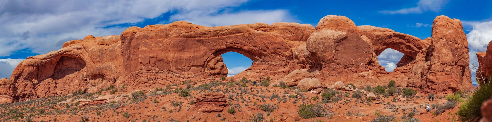 Panoramic view of North Window, South Window, and Turret Arch with beautiful clouds in the sky, small figures of people in the frame emphasize the scale and beauty of this majestic creation, Moab, USA