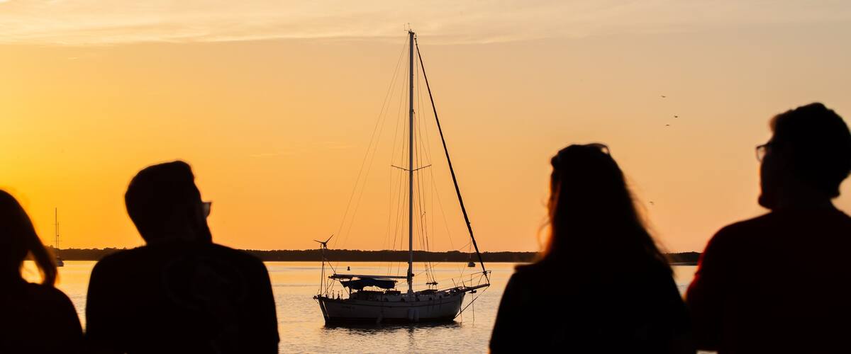 Silhouettes of people at sunset, a sailboat on the water, and a distant shoreline.