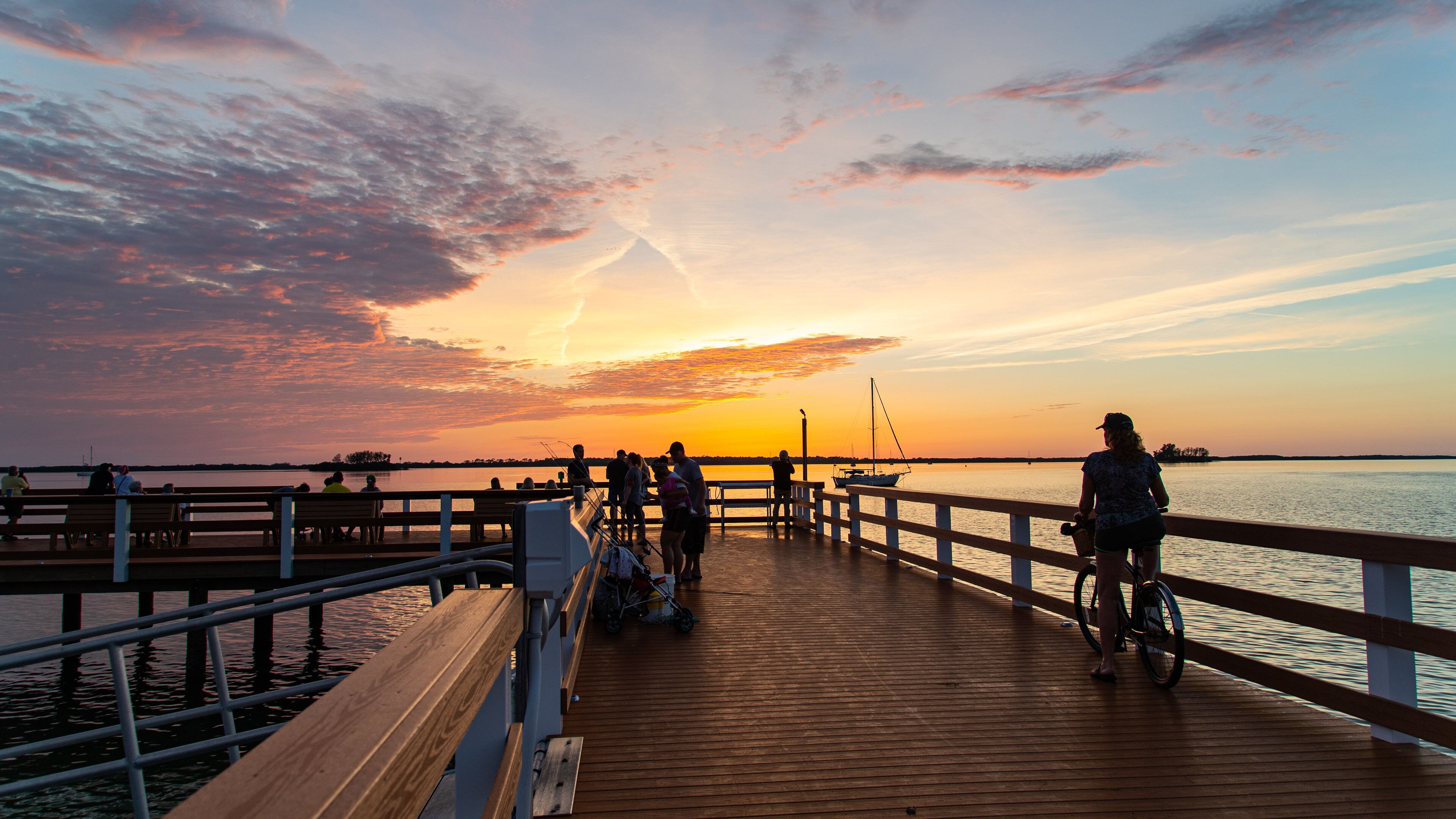 A wooden pier extending into a body of water during sunset with people and a sailboat visible.