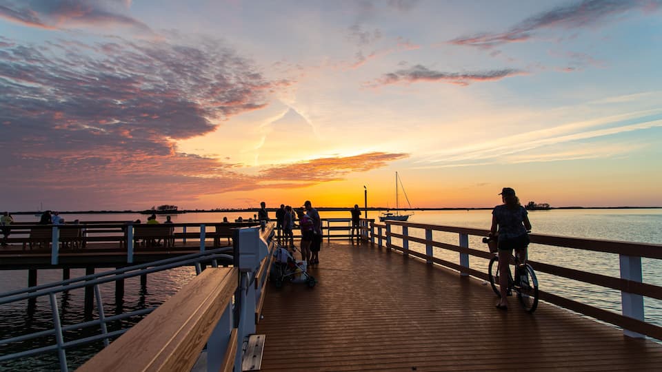 A wooden pier extending into a body of water during sunset with people and a sailboat visible.