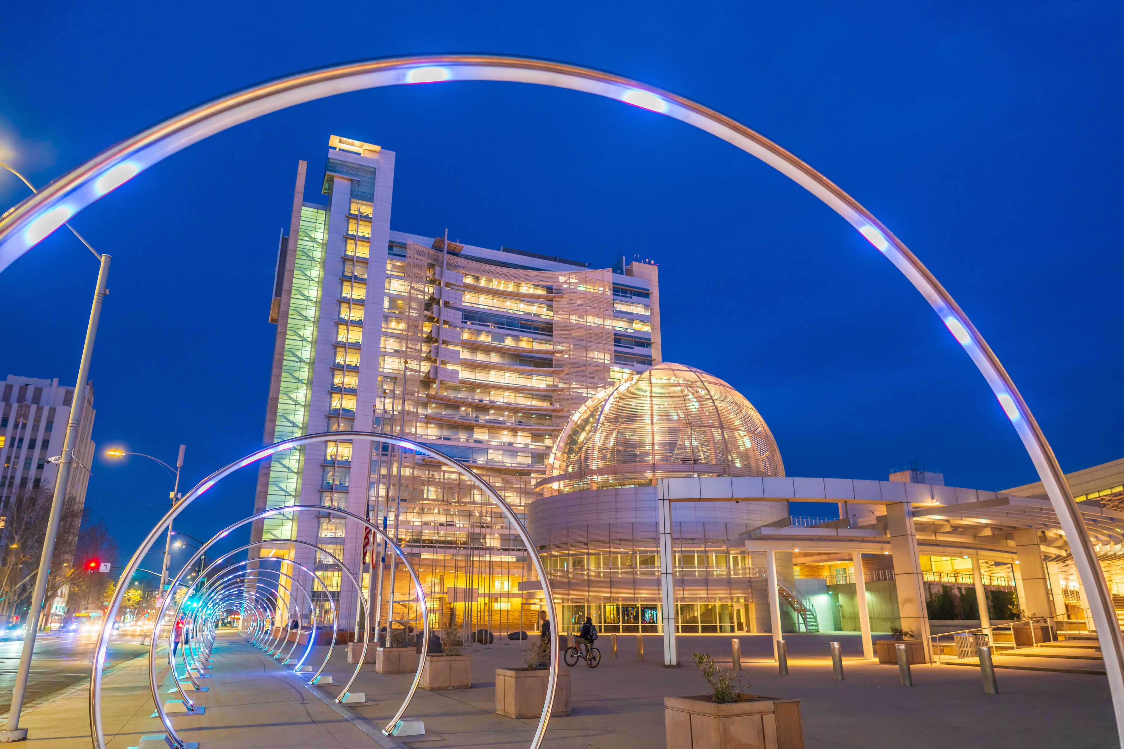 Een modern gebouw met een koepel en een gebogen glazen gevel, omgeven door een voetgangersgebied met een lichtinstallatie.