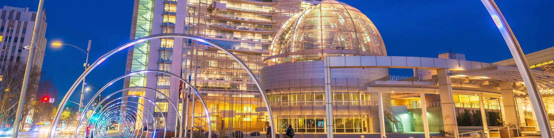 Un edificio moderno con una cúpula y fachada de vidrio curva, rodeado de una zona peatonal con una instalación de luces.
