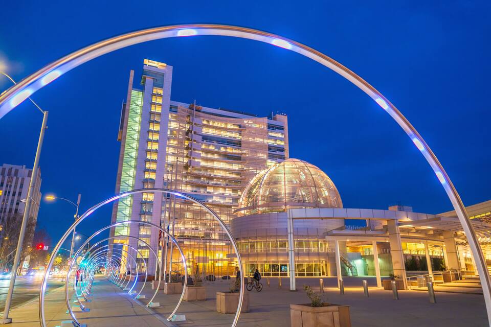 Een modern gebouw met een koepel en een gebogen glazen gevel, omgeven door een voetgangersgebied met een lichtinstallatie.