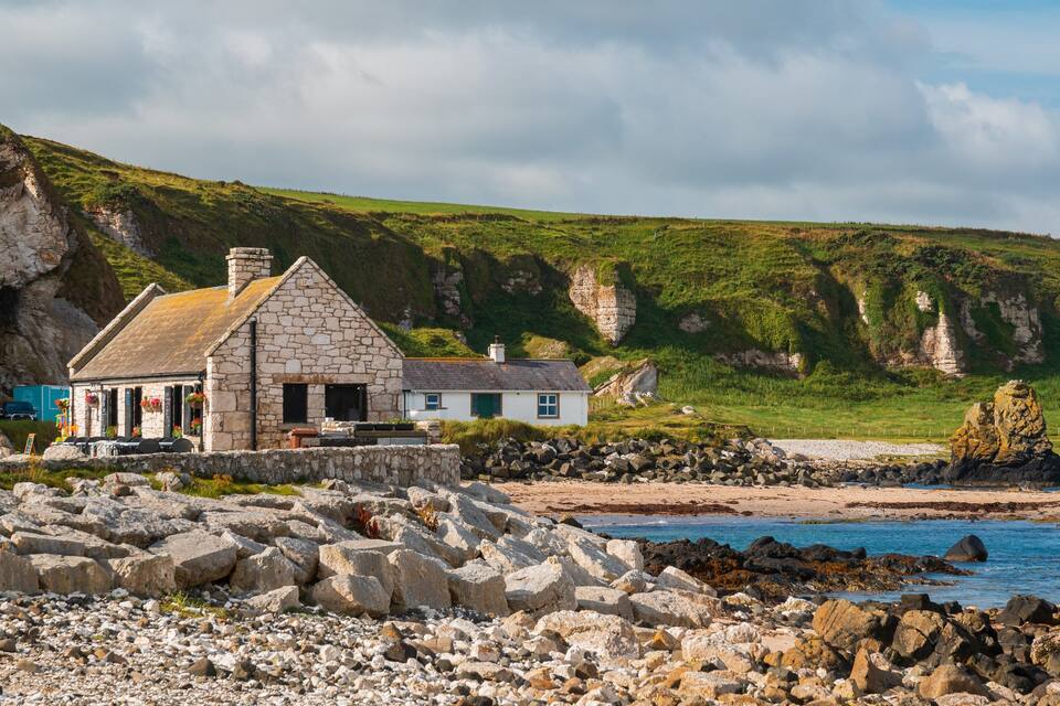 A stone house by a rocky beach with cliffs in the background.
