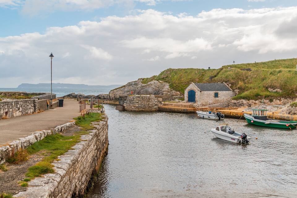 A stone pier with boats, a small stone building, and a grassy hill with a windmill.