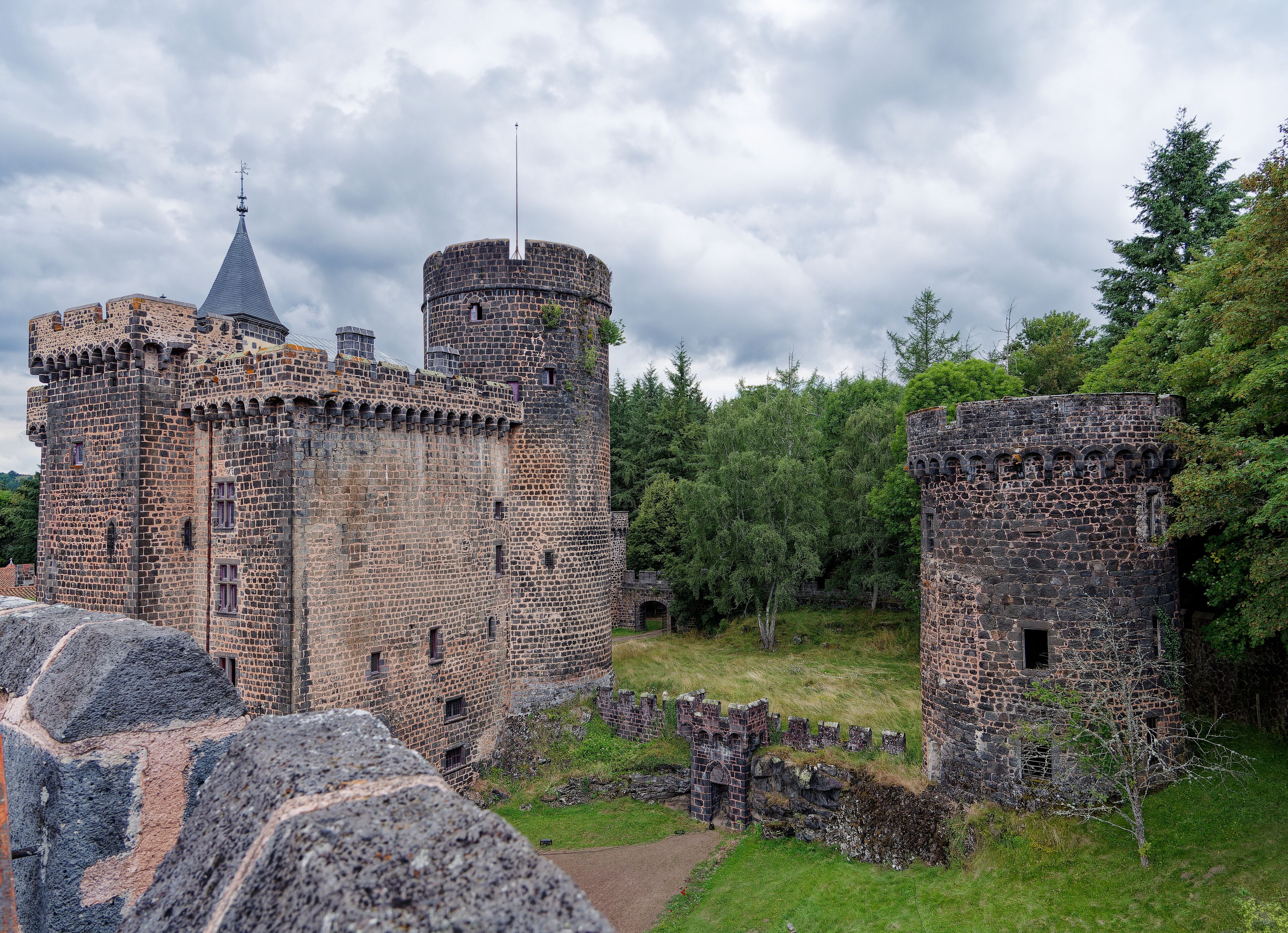 A historic stone castle with two towers, surrounded by lush greenery and a cloudy sky.