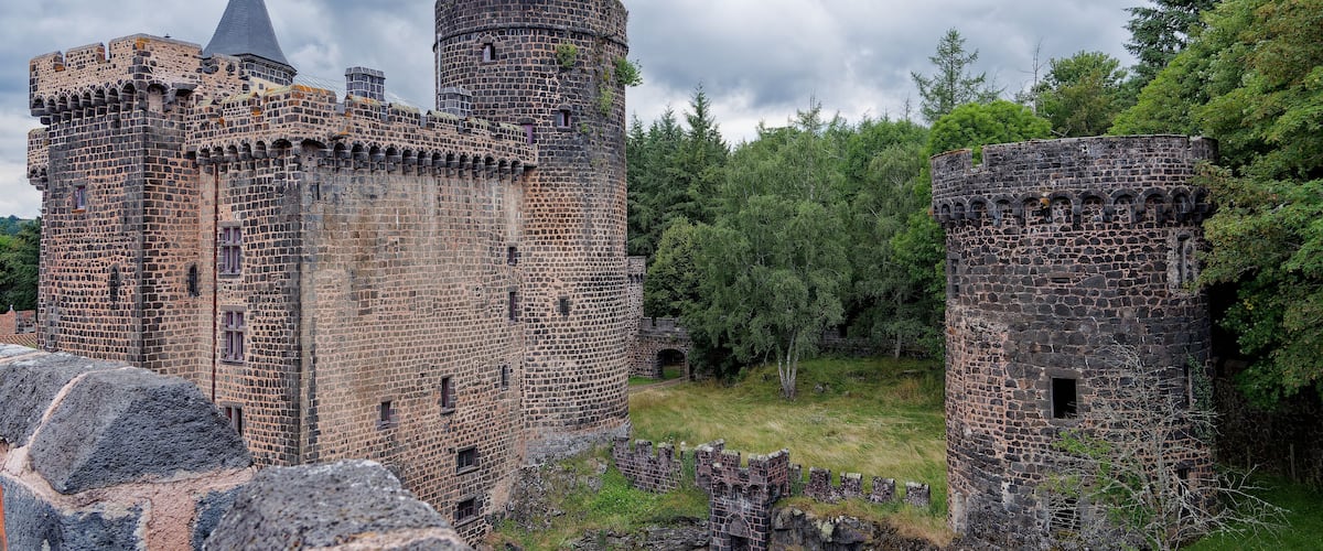 A historic stone castle with two towers, surrounded by lush greenery and a cloudy sky.