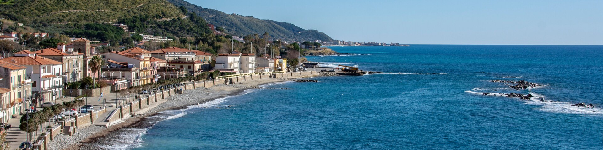 Un pueblo costero con edificios a lo largo de la costa, un rompeolas rocoso y un mar azul y transparente.