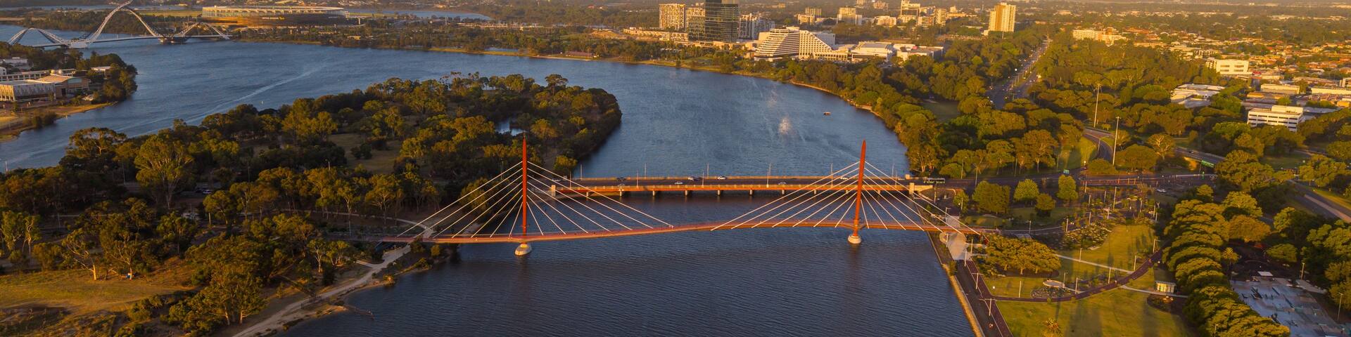 A large suspension bridge over a river, surrounded by greenery and urban buildings.