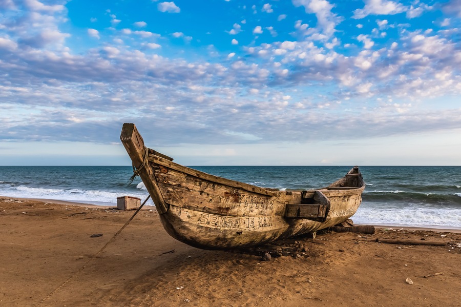 Um barco desgastado numa praia arenosa, com o oceano ao fundo.