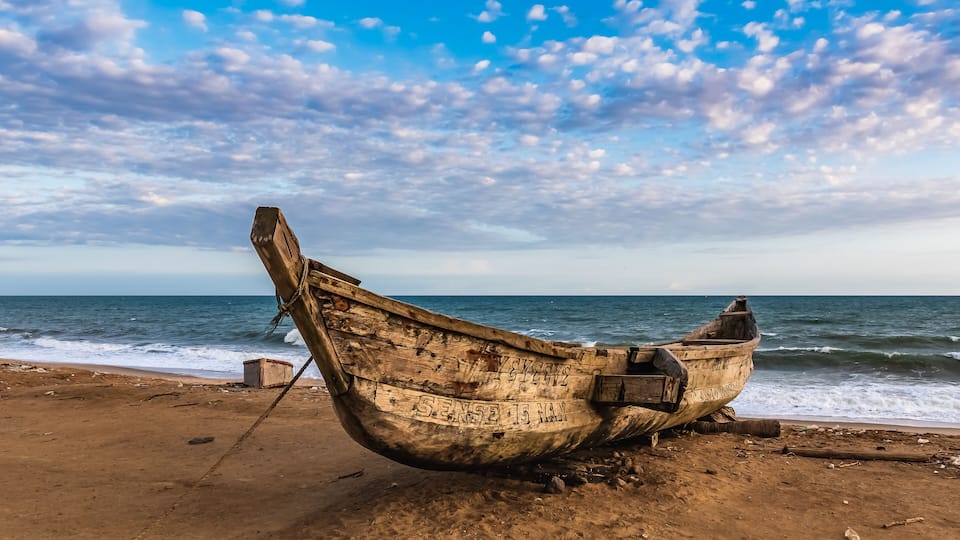 A weathered boat on a sandy beach with the ocean in the background.