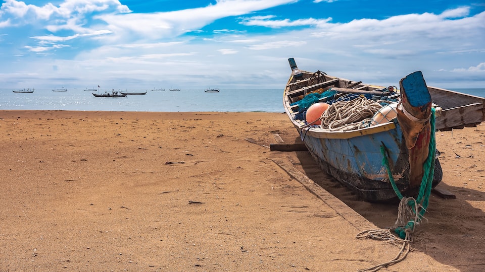A beach with a weathered boat tied to a wooden post, the ocean in the background with several boats visible.
