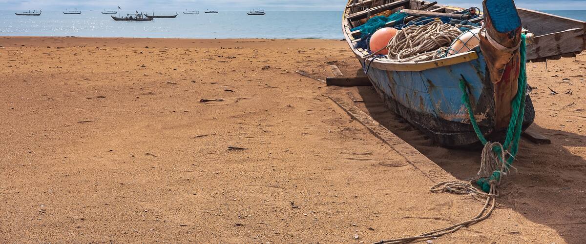 A beach with a weathered boat tied to a wooden post, the ocean in the background with several boats visible.