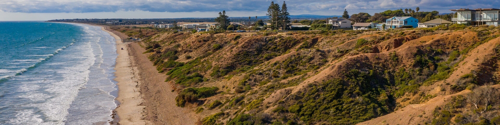 Un paisaje costero con una playa arenosa, colinas onduladas y casas al fondo.