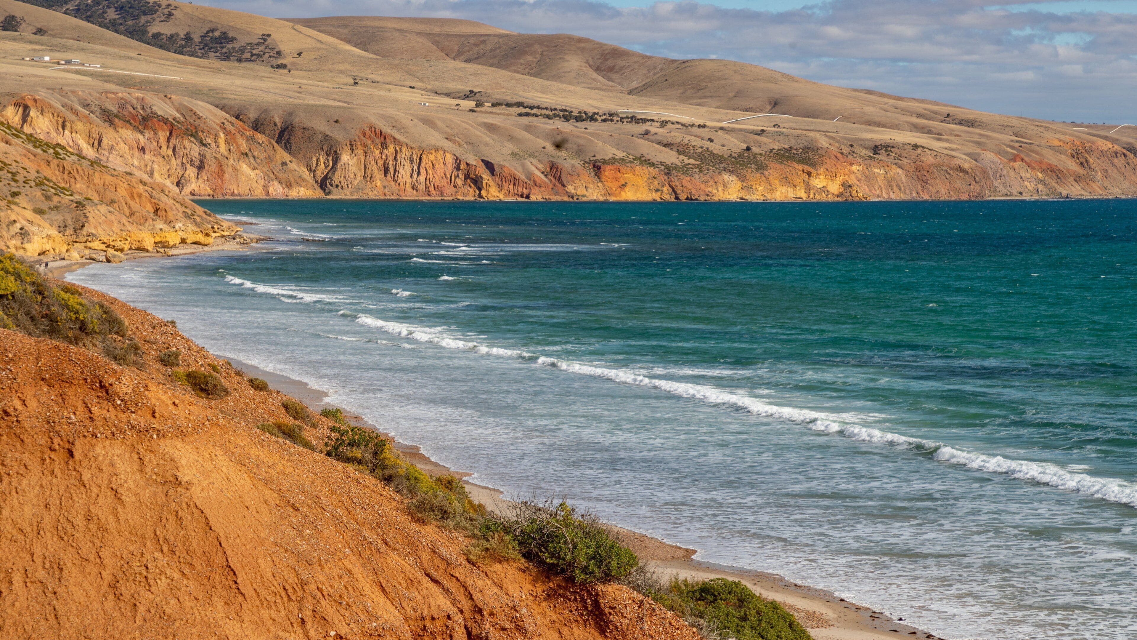 Un paisaje costero con una playa arenosa, olas oceánicas y colinas ondulantes al fondo.