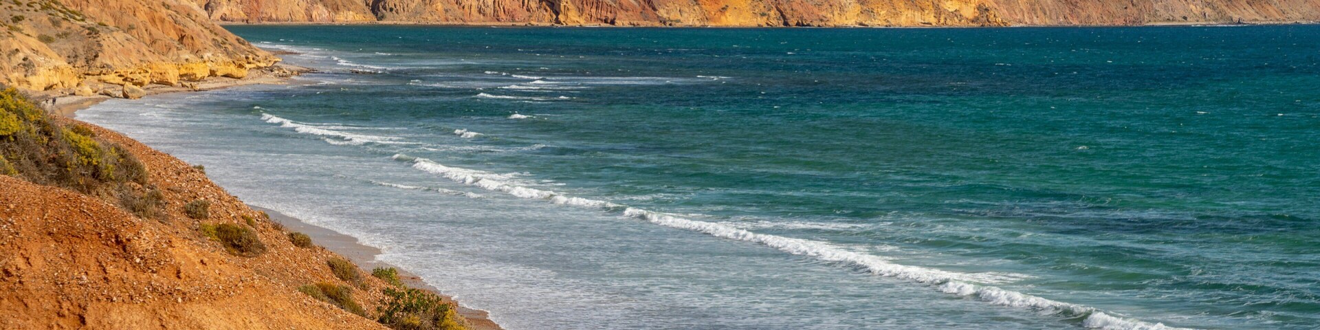 Un paisaje costero con una playa arenosa, olas oceánicas y colinas ondulantes al fondo.