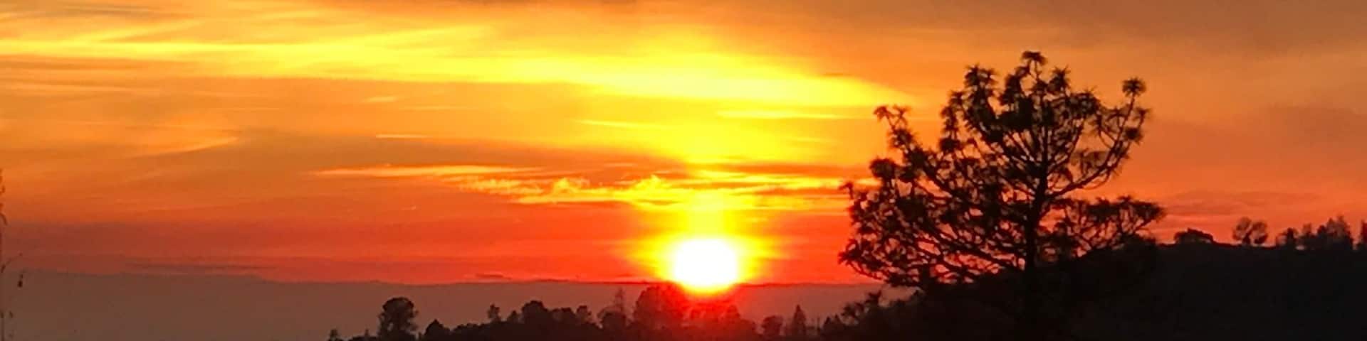 A sunset over a hilly landscape with silhouettes of trees and a clear sky.