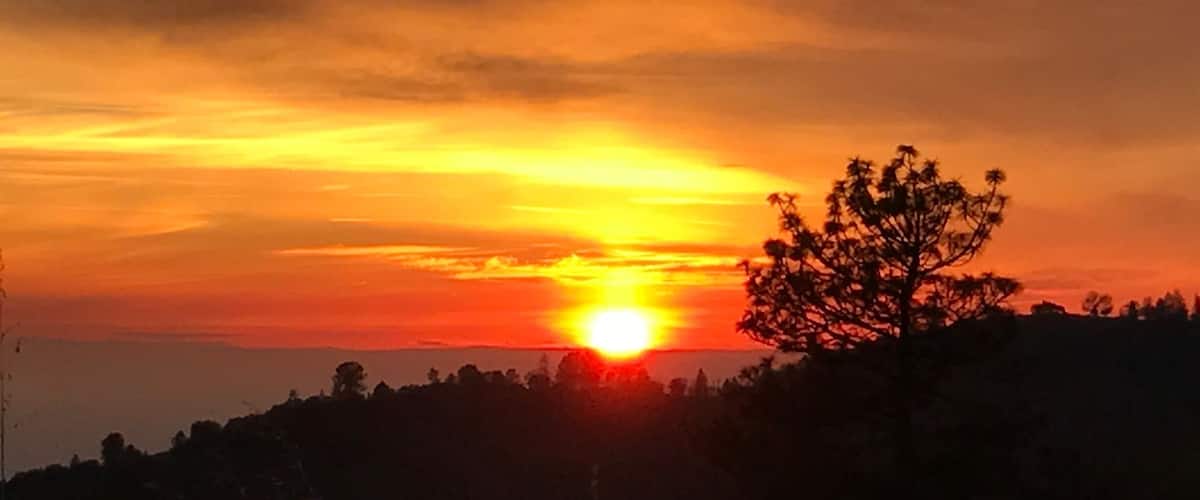 A sunset over a hilly landscape with silhouettes of trees and a clear sky.